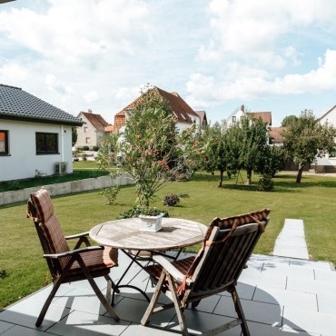 Ein Tisch und zwei Stühle stehen auf einer gepflasterten Terrasse in einem großen Garten auf einem größeren Grundstück hinter dem Haus.
