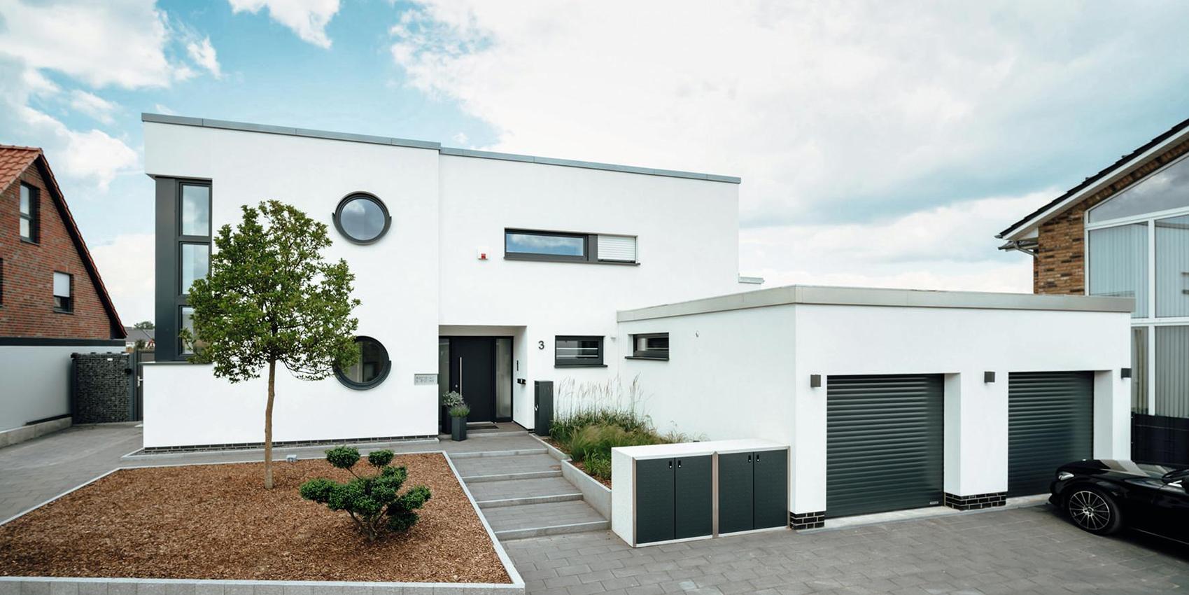 Im Vordergrund steht ein modernes rechteckiges Haus mit Flachdach, während im Hintergrund und nebenan typischere Häuser mit Schrägdächern zu sehen sind.