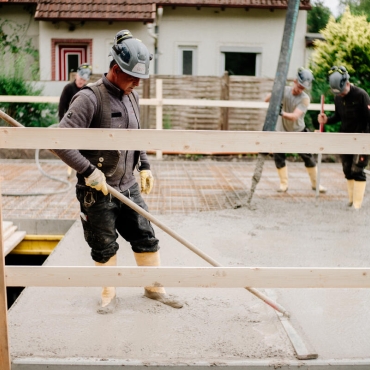Ein Bauarbeiter im Vordergrund glättet den Beton im Rahmen des Hausbaus. Drei weitere Bauarbeiter sind im Hintergrund zu sehen.