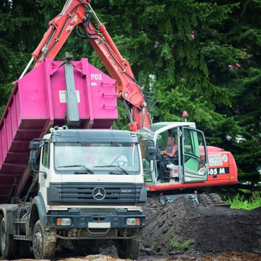 Ein Baufahrzeug gräbt Erde aus, die von einem Kippwagen abtransportiert wird.