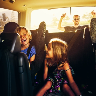 Eine Familie ist unterwegs, drei Kinder sitzen auf dem Rücksitz des Fahrzeugs und der Vater schaut durch das Rückfenster zu.